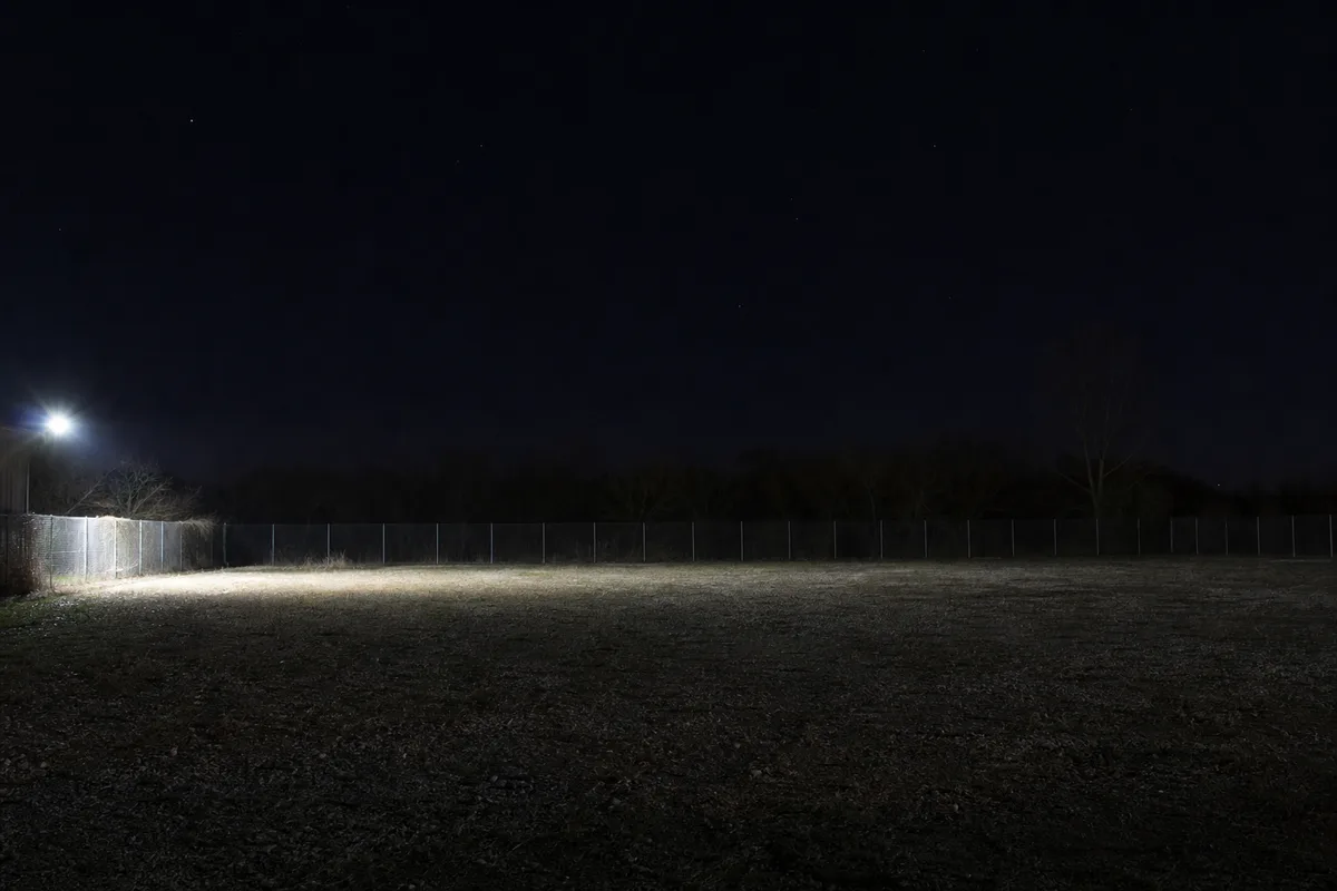 Terrain vide clôturé de nuit, éclairé par un unique projecteur